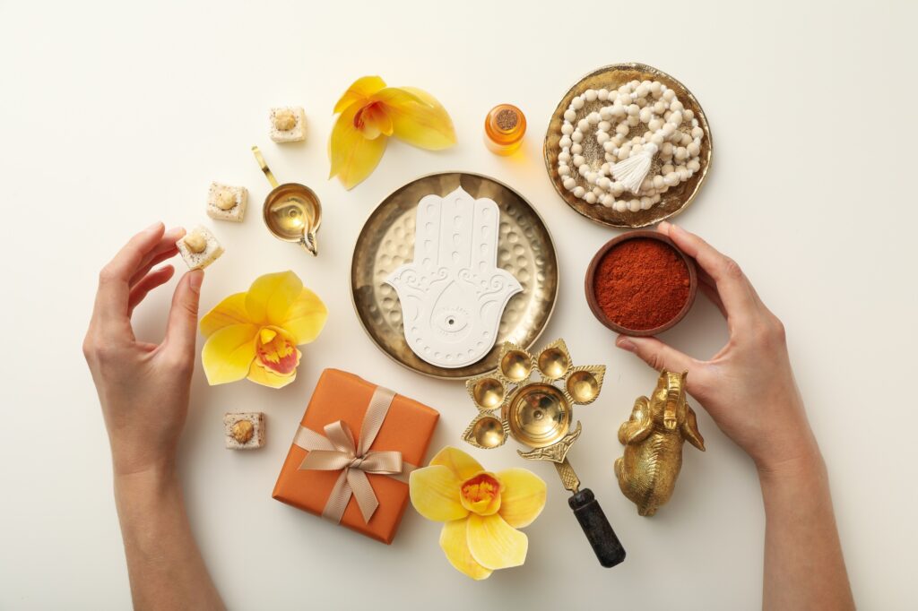 Flat lay of spiritual items including a mala bead necklace, symbolic Hamsa hand, sacred powder, flowers, sweets, and decorative objects associated with rituals and jewelry practices.