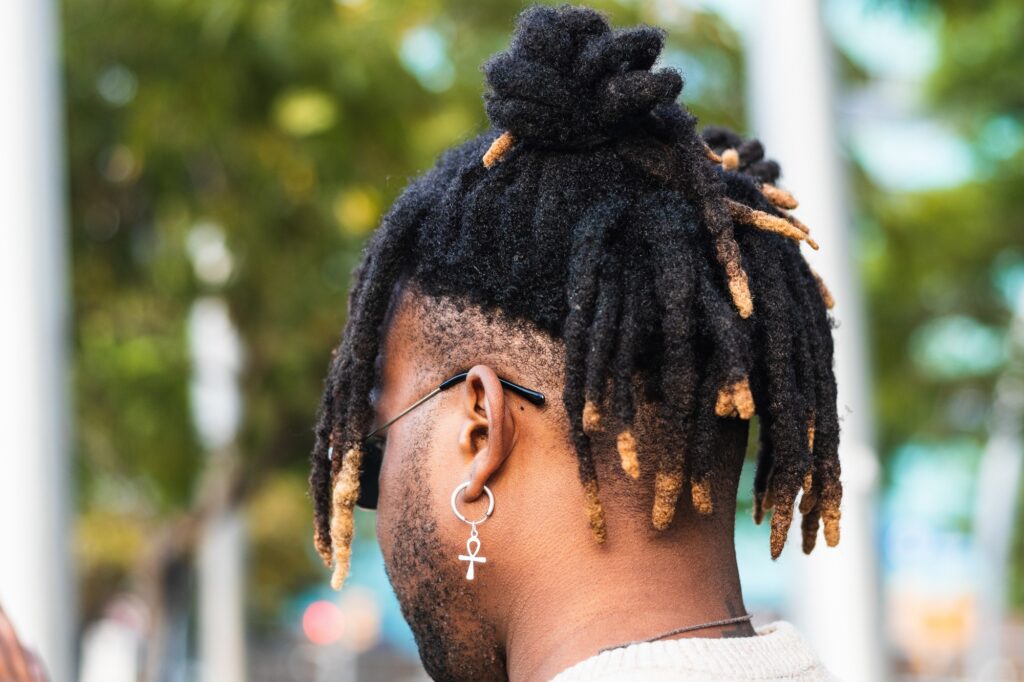 Close-up of a person with locs and an ankh symbol earring, viewed from behind, with natural outdoor background.