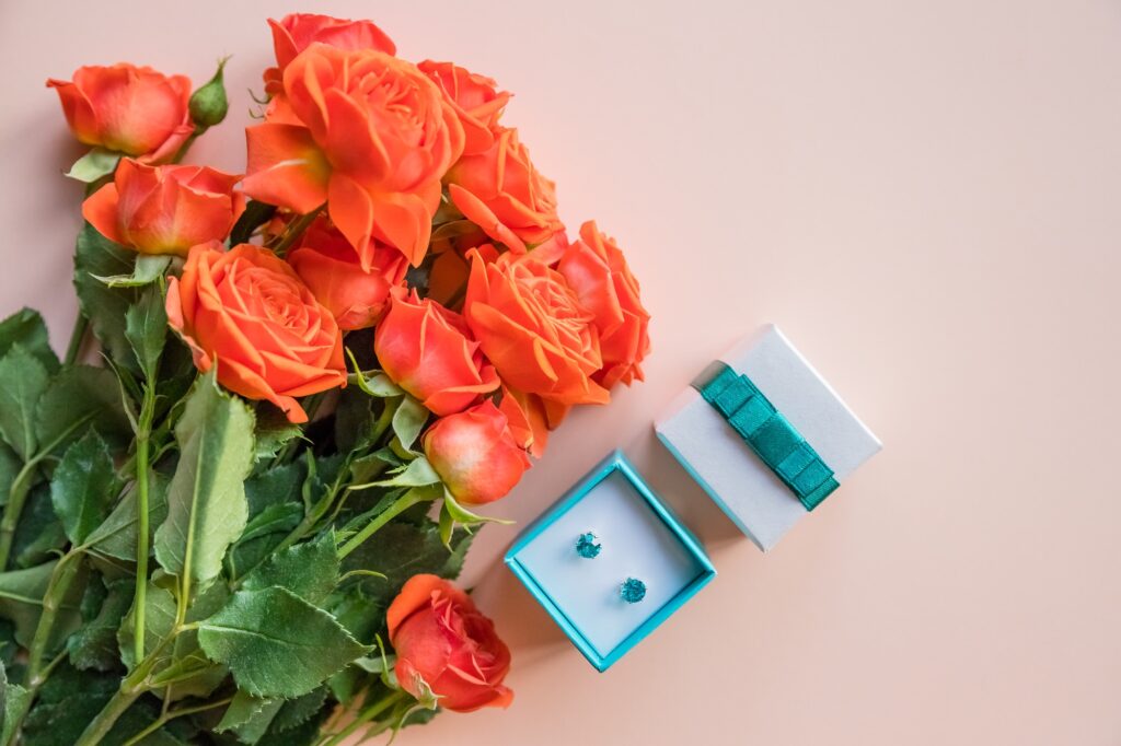 A bouquet of vibrant orange roses next to an open gift box containing turquoise stud earrings, with a matching closed gift box beside it, all set against a soft peach background.